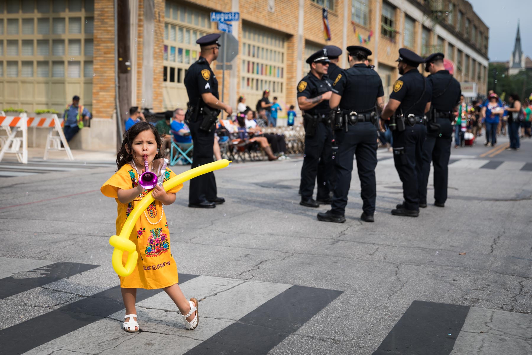 Little girl in yellow dress blowing toy horn and holding yellow balloon in front of cops for an article on the Fiesta photo tips