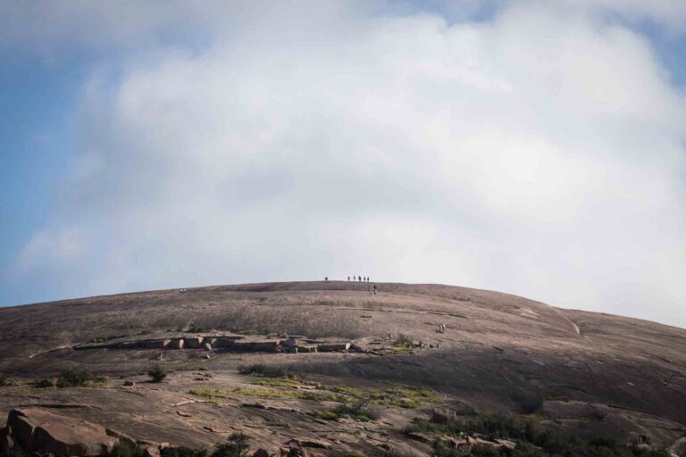 Small speck of people on top of mountain with clouds overhead for an article on Enchanted Rock hiking trips