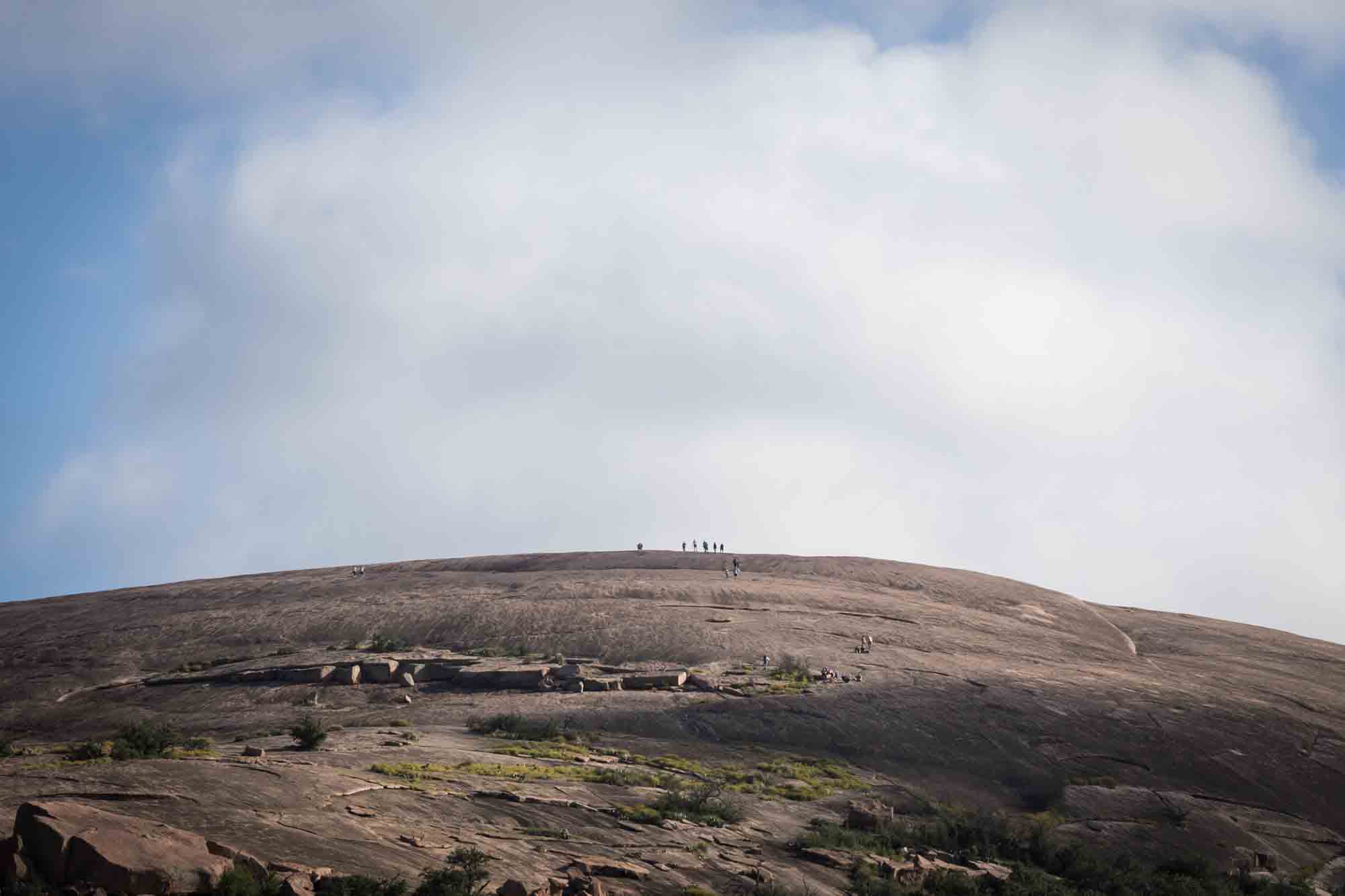 Small speck of people on top of mountain with clouds overhead for an article on Enchanted Rock hiking trips