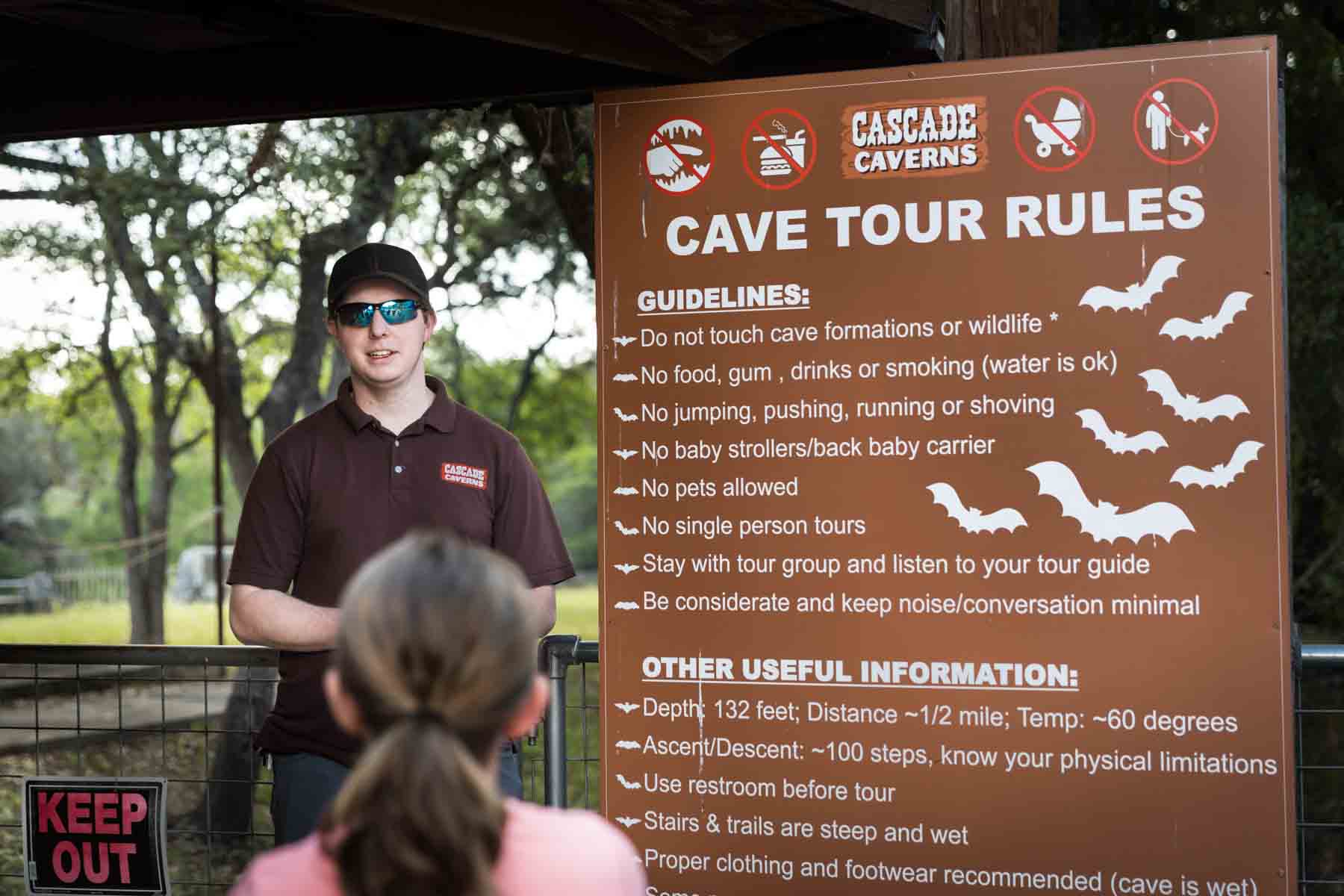 Tour guide wearing sunglasses beside sign in Cascade Caverns for an article entitled, ‘Visiting Boerne, Texas: Everything You Need to Know’